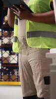 Vertical African american female staff in hi vis vest counting packages on industrial racks, processing custom orders for e-commerce retail operations. Large business management in storehouse. Camera A. video