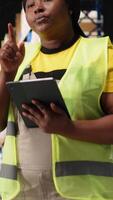 Vertical African american female staff in hi vis vest counting packages on industrial racks, processing custom orders for e-commerce retail operations. Large business management in storehouse. Camera B. video