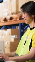 Vertical Two colleagues working on order processing in industrial warehouse space, wearing hi vis vests near huge storage racks. Asian man typing shipment details on the computer. Camera B. video