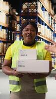 Vertical African american and asian team presenting the cargo goods on a live stream , recording a vlog in industrial warehouse near racks. Content for large business advertisement. Camera A. video