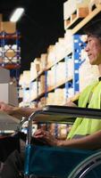 Vertical Asian man wheelchair user typing tracking numbers on a computer software, updating parcel status in real time. Staff managing order fulfillment in a logistics center to sort inventory. Camera A. video