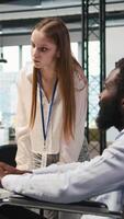 Vertical Junior dev and coworker in wheelchair developing software applications, optimizing code. Female technician and colleague with paraplegia in office using AI to solve bottlenecks, camera A video