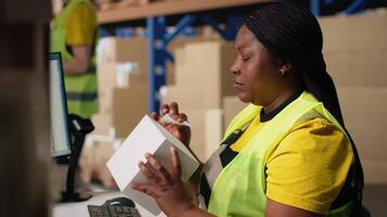 African american woman printing and applying airway bill tags on boxes, wearing hi vis vest in a warehouse near racks. Managing barcode labels with tracking info for order fulfillment. Camera B. video