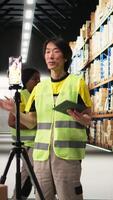 Vertical Asian worker promoting the large business industrial warehouse in a vlog, live streaming a for e-commerce shipment advertisement. Staff presenting the storage on commercial ad. Camera A. video