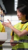 Vertical African american and asian team presenting the cargo goods on a live stream , recording a vlog in industrial warehouse near racks. Content for large business advertisement. Camera B. video