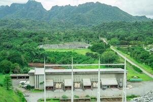 Hydropower plant building with green forest and high-voltage infrastructure, with mountain landscape in background. Hydroelectric power station at dam site. Renewable energy. Power generation. photo