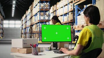 Logistik Arbeiter verwaltet Aufträge Nächster zu Grün Bildschirm Anzeige, präsentieren Warenhaus Automatisierung Werkzeuge und Inventar Steuerung Dashboards. E-Commerce bestellen wird bearbeitet mit Lager Gestelle. Kamera a. video