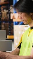 Vertical Man in hi vis vest reviews shipment details on awb tags for dispatch in industrial warehouse, parcels from the storage racks with tracking numbers for seamless express delivery. Camera B. video