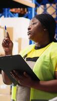 Vertical Black female worker verifying awb labels on files for inventory control, counting packages on racks and ensuring fast delivery in the warehouse. E-commerce airway bills tags. Camera B. video