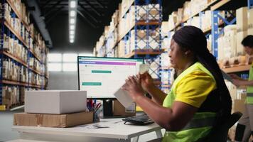 Black warehouse employee manage labeling and packaging tasks, applying printed airway bill tracking numbers tags on packages. Woman in hi vis vest processing orders in stock room. Camera A. video