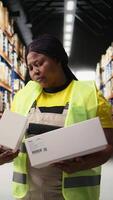 Vertical Portrait of african american worker checking packed boxes and labels before cargo shipment, ensuring order fulfillment for retail online shopping large business. Logistics center activity. Camera A. video