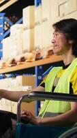 Vertical Asian man in wheelchair processing orders and preparing the boxes for distribution in fulfillment center, wears hi vis vest in e commerce logistics facility. Retail fast delivery. Camera A. video