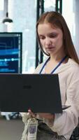 Vertical Portrait of smiling female engineer coding with AI language model assistance in startup office, using notebook. Happy young worker typing on laptop, using LLM assistant, camera A video