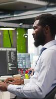 Vertical Engineer coding with AI language model assistance in startup office, listening music. African american man wearing headphones integrating LLM assistant suggestions into code editor, camera A video