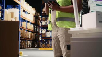 African american female staff in hi vis vest counting packages on industrial racks, processing custom orders for e-commerce retail operations. Large business management in storehouse. Camera A. video