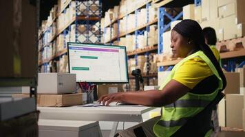 African american employee updating the order logistics software, preparing the shipment labels for distribution. Industrial depot staff with hi vis vest in e commerce logistics facility. Camera A. video