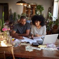 A man and woman sit at a table with money and a laptop photo