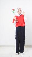 Beautiful Asian girl celebrate Indonesia independence day in red and white outfit holding a rose and showing winning pose with raising her clenched fist isolated on white background photo