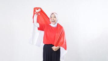 Happy Asian girl celebrate Indonesia independence day in red and white outfit showing victory pose with Indonesian flag draped around her back like a cape isolated on white background photo