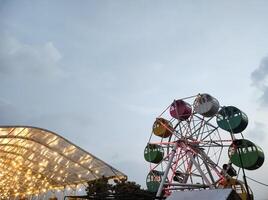 A mini Ferris wheel with a sunset sky in the background. The gondolas, or cabins, of the Ferris wheel are brightly colored. An amusement ride on the rooftop of a shopping center. Negative space. photo