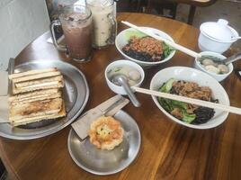 Tabletop shot of a typical Indonesian lunch menu. The dish consists of chicken noodles with meatball soup, a cold chocolate drink, and desserts of toast and egg tart. Indonesian gourmet photo