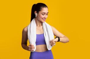 Sport And Technology Concept. Smiling Young Woman With Towel On Neck Looking At Wearable Bracelet, Checking Activity Tracker, Wearing Wireless Earbuds Isolated On Orange Studio Background photo