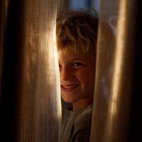 Playful Child Peeking Through Curtains in Warm Light. A charming close up portrait of a boy hiding behind sheer curtains, illuminated by soft sunlight. photo