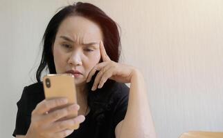 Depressed mature Asian woman sit and looking on phone, lost in thoughts, thinking about problem. photo