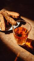 A glass of amber liquid sits next to a plate of biscotti on a wooden table, bathed in warm sunlight. photo