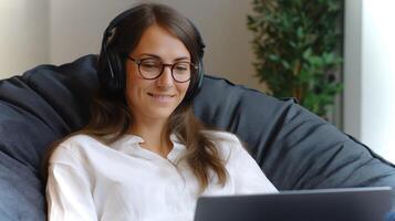In a cozy setting, a young woman sits in a bean bag, wearing headphones and smiling as she engages with her laptop, showing concentration and joy in her task. photo