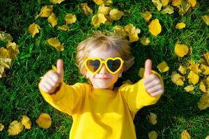 Young Boy in Red Sunglasses Gives a Double Thumbs Up while lying on vibrant green grass scattered with yellow leaves. photo