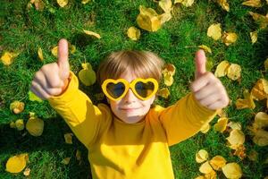 A Young Boy in Red Sunglasses Gives a Double Thumbs Up while lying on vibrant green grass scattered with yellow leaves. photo