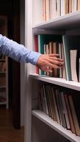 vertical Close-up of a male hand browsing through books on a white bookshelf in a library or bookstore, searching for information or leisure reading video