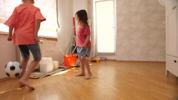 Two kids enjoy a game of soccer inside a bright room, skillfully maneuvering a black and white ball on a polished wooden floor. Sunlight filters through a nearby window, adding warmth video