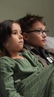 vertical Brother and sister sitting on a sofa, watching television with expressions of boredom, clearly showing disinterest in the program playing on the screen video
