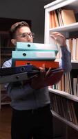 vertical Stressed office worker carrying a heavy stack of colorful binders while navigating through a library or archive, surrounded by shelves filled with books and documents video