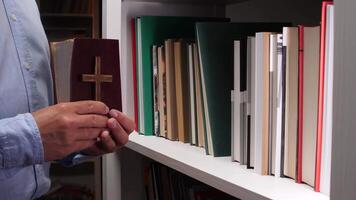 Male hands holding a bible alongside a wooden cross, surrounded by a bookshelf filled with books, embodying a deep sense of faith and spirituality in a serene indoor setting video