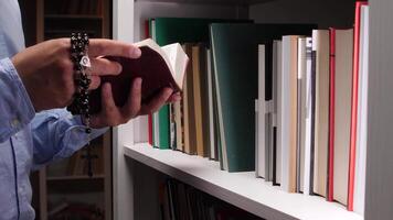 Catholic priest wearing a blue shirt, reading a holy book and holding rosary beads, stands amidst shelves filled with books in a serene library setting, embodying faith and devotion video