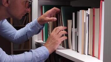 Male librarian browsing carefully through books on a white bookshelf, searching intently for a specific title among the vast collection in a library or bookstore setting video