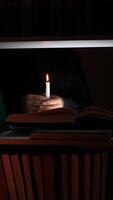 Hand holding a burning white candle, casting a warm glow across a bookshelf filled with books in a dark room, creating an inviting and mysterious ambiance perfect for quiet reading video