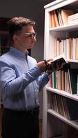 vertical Young male believer wearing glasses is standing next to a white bookshelf in a library, reading a holy book and holding rosary beads, searching for spiritual guidance and praying video