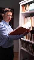 vertical Office worker placing an orange folder on a high shelf in a white bookcase at night, surrounded by numerous books, focusing on organization and tidiness in the workspace video