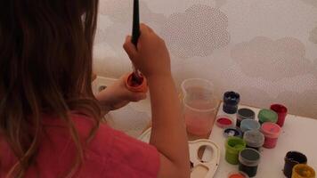 Young girl preparing for a painting session, dipping her brush into a vibrant orange paint pot, surrounded by an array of colorful supplies and water, fully immersed in her creative activity video