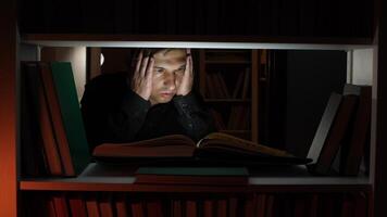 Focused student is engrossed in a book, illuminated by a small light within a dark library, holding his head, suggesting deep concentration or fatigue during a late-night study session video