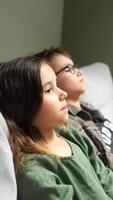vertical Brother and sister sitting on a sofa, watching television with expressions of boredom, clearly showing disinterest in the program playing on screen video