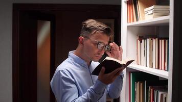Young man wearing glasses rubbing his tired eyes while reading a book and standing next to a bookshelf in a library, experiencing eye strain from prolonged studying video