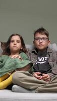 vertical Bored brother and sister are sitting on a comfortable sofa, watching television and holding a remote control in their hands in a cozy living room video