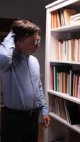 vertical Pensive librarian wearing glasses scratches his head while searching for a specific book, surrounded by shelves filled with literature in a library or bookstore video