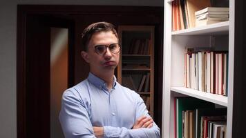 Portrait of a serious male teacher or librarian wearing glasses and a blue shirt, standing with crossed arms in a library or bookstore, looking confident and professional video