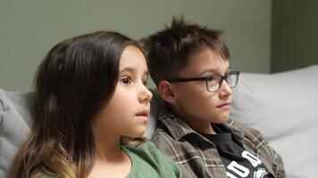 Brother and sister sitting on a sofa, watching television with expressions of boredom, clearly showing disinterest in the program playing on the screen video